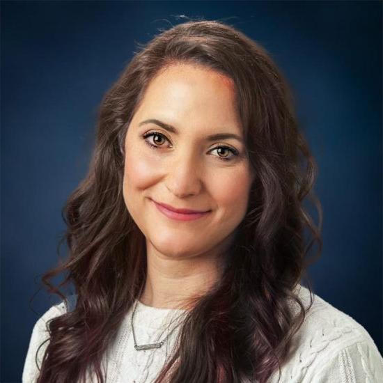 Professional portrait of a woman with brown hair, smiling and wearing a white sweater with a necklace, set against a dark blue background.